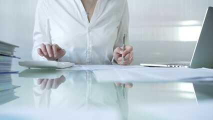 Businesswoman in blue blouse is using white calculator and taking notes while working on financial...