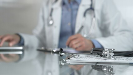 Stethoscope and medication history records form are on a glass table before a male doctor working with laptop in clinical office background. Medicine concept