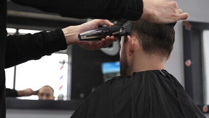 Professional barber trimming male client's hair using electric razor, creating contemporary...