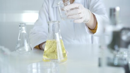 A scientist, wearing a lab coat and white protective gloves, is pouring a yellow oily liquid from one beaker to another in laboratory, close up. Medicine and science concept