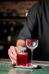A bartender presents a red cocktail in a stylish glass garnished with a heart-shaped ice cube, alongside a decorative bottle on a green bar countertop.