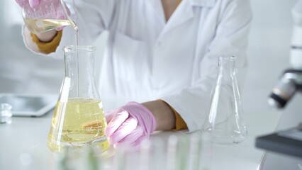 A scientist, wearing a lab coat and pink protective gloves, is pouring a yellow oily liquid from one beaker to another near microscope in laboratory, close up. Medicine and science