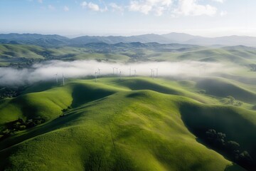 Obraz premium Aerial view of green rolling hills with fog in the valley and wind turbines on the horizon in a serene landscape under a clear blue sky.