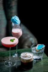 A vibrant display of cocktails featuring a pink drink garnished with mint, a cotton candy-topped beverage, a classic whiskey drink, and an oyster with blue detail, set against a dark background.