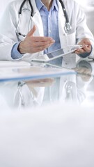 Unknown male doctor wearing a stethoscope, blue shirt and a lab coat is gesturing with hands overs a glass desk with digital tablet in clinic office, vertical closeup view. Medicine concept