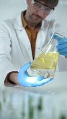 Man scientist, wearing blue gloves and protective glasses, is holding a yellow chemical solution...