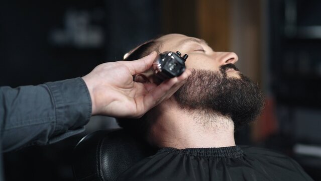 Professional barber trimming client's beard with electric razor, delivering precise styling within contemporary grooming salon setting