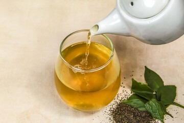 The teapot pour green tea into a glass on the table. Herbal tea.