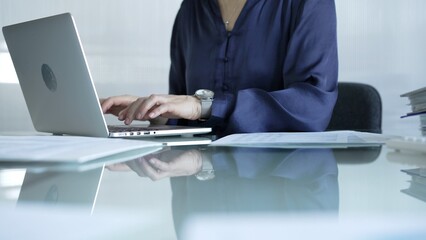 Businesswoman in blue blouse typing on laptop at glass desk while sitting in bright office setting, close-up vie. Businesspeople concept