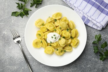 Tasty dumplings with green onion, sour cream and fork on grey textured table, flat lay