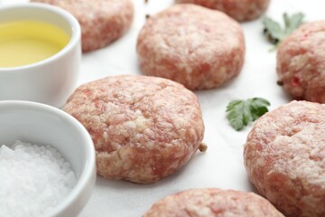 Many uncooked patties, salt, oil and parsley on white table, closeup