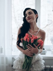 Sensual sexy woman in bridal erotic bodysuit posing against window with bouquet of red tulips in...