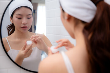 Fototapeta premium Beautiful young asian woman applying cream or lotion for rejuvenation skin on face while looking mirror in bathroom, woman applying moisturizer for treatment wrinkles on facial, beauty and skincare.