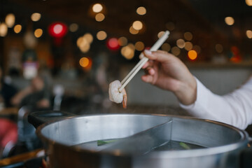 Close-up of a hand holding shrimp with chopsticks over a steaming hot pot. Traditional Asian shabu-shabu or hotpot dining experience in a cozy restaurant. Perfect for food, and lifestyle themes.