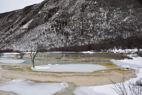 Scenic winter landscape of snow-covered mountains, partially frozen turquoise pools, and bare trees creating a serene and picturesque view. 