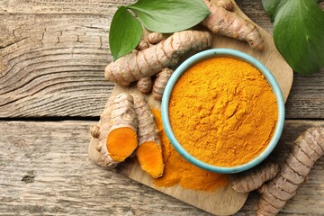 Turmeric powder in bowl, roots and leaves on wooden table, flat lay. Space for text
