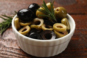 Different delicious marinated olive rings, whole ones and rosemary in bowl on wooden table, closeup