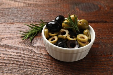 Different delicious marinated olive rings, whole ones and rosemary in bowl on wooden table, closeup