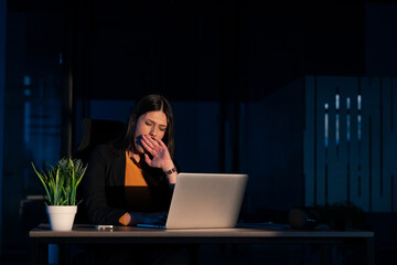 Tired businesswoman working late at night, yawning while sitting at a desk with a laptop in a dimly lit office. Concept of work fatigue, overtime, and exhaustion in a corporate setting