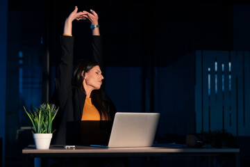 Young female entrepreneur or executive stretches and yawns while working late at night in a dimly lit office, showing signs of fatigue. Concept of overworking, exhaustion, long hours at the workplace