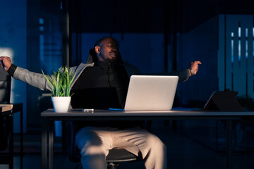Tired African businessman stretching at his desk in a dimly lit office, taking a break from work....