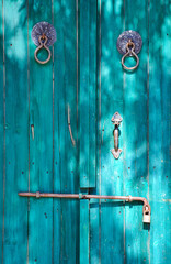 Close-up of a vintage turquoise wooden door with rustic handle and locks, showing weathered textures and shadows. Ideal for themes of historical architecture, rustic charm, and old-world aesthetics.