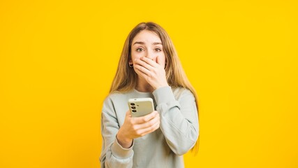 Shocked young woman covering her mouth while reading news on smartphone on yellow background