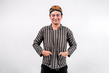 A young Indonesian man wearing traditional sorjan or lurik attire stands confidently with a smile, hands in his pockets, showcasing a blend of cultural heritage and modern style on neutral background.