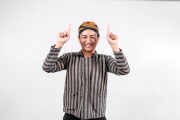 A young man wearing traditional lurik attire smiles brightly while pointing upwards with both...