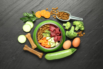 Natural and dry pet food in feeding bowl among products on dark textured table, flat lay