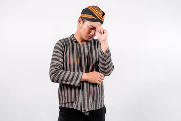 A young man wearing traditional lurik attire expresses frustration while resting his hand on his forehead, showcasing cultural pride and emotion against a neutral background.