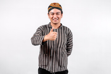 A young man wearing traditional lurik attire smiles broadly while giving a thumbs-up gesture, showcasing cultural pride and positivity against a neutral background.