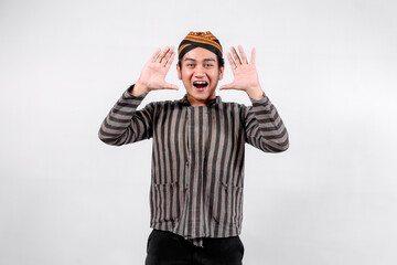 A young man wearing traditional lurik attire smiles widely while making a joyful gesture with both hands, showcasing cultural pride and enthusiasm against a neutral background.