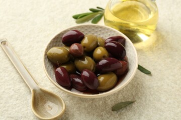 Tasty marinated olives in bowl, leaves and oil on beige table, closeup
