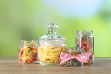 Tasty gummy candies in jars on wooden table against blurred green background