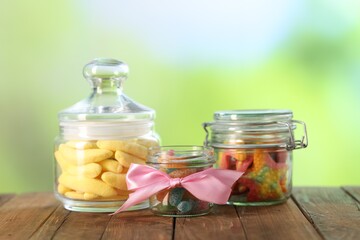 Tasty gummy candies in jars on wooden table against blurred green background