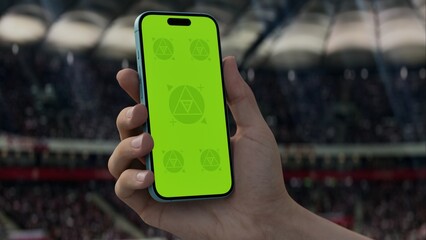 GREEN SCREEN CHROMA KEY A hand holding a phone with a green screen at a stadium during a football match. Ideal mockup for sports, betting, or event apps
