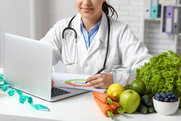 Young female nutritionist with laptop and healthy food writing diet plan in office, closeup