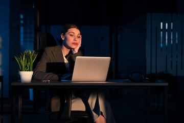 Tired businesswoman working late at night in a dimly lit office, staring at a laptop with a...