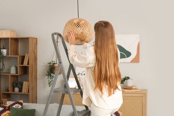 Young woman screwing light bulb into ceiling lamp on stepladder at home, back view