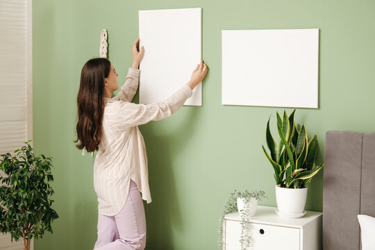 Young woman hanging empty canvas on green wall in bedroom