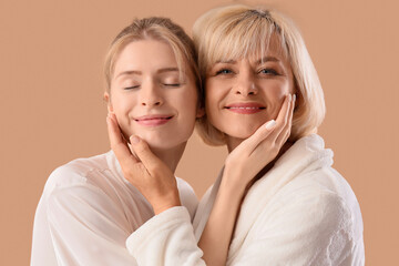 Happy young woman and her mother in bathrobes hugging on beige background