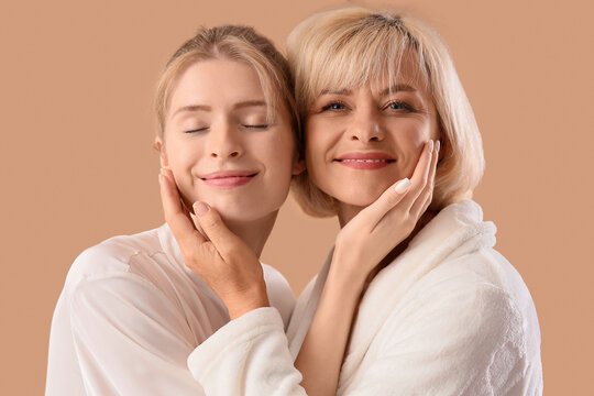 Happy young woman and her mother in bathrobes hugging on beige background