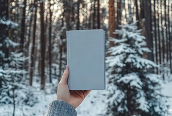 Winter reading retreat. Hand holding light blue book against snowy forest background, perfect for contemplative winter lifestyle and outdoor reading concepts.