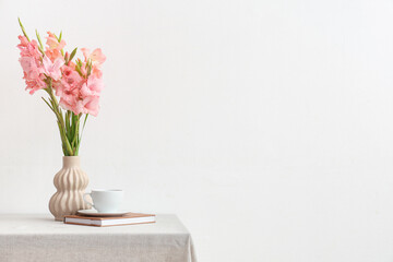 Vase with bouquet of beautiful gladiolus flowers, book and cup of coffee on table against white...