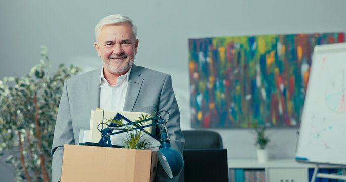 Retirement of experienced long-time director, manager, head of the company, older man with gray hair holds a cardboard box with packed things in hands, looks inside, smiles, rest, vacation, retired