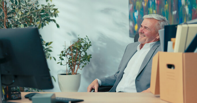 Smiling senior company executive sits in comfortable swivel chair next to computer looking around him reminiscing about years of work elegant man with gray hair spends last day before retirement