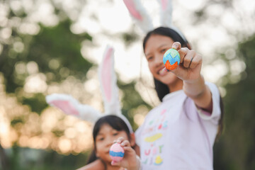 Obraz premium Focus of Easter Egg in the hand of The girl wear cute bunny ears on the head on background of a natural garden with sunset.