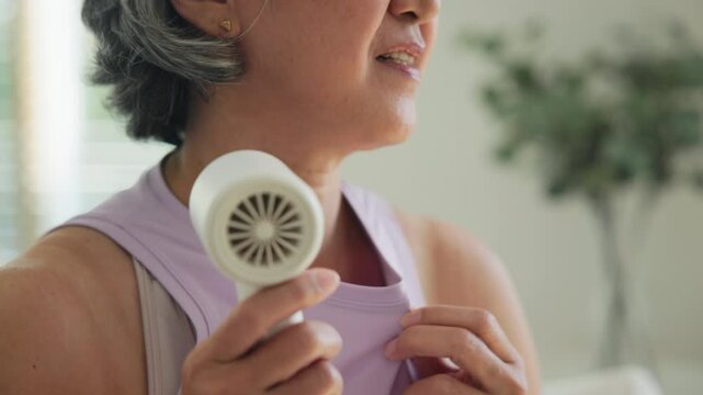 Asia adult people middle age old woman sit at sofa home feel hot flash fever use fan waving hand blowing wind face elder health care summer heat aging issue. Sad pain sweat tired stroke suffer stress.