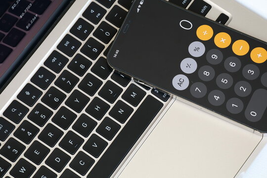 A sleek gold laptop with a smartphone displaying a calculator app rests on a white surface. The laptop screen shows cryptocurrency market data, creating a modern financial and tech-focused atmosphere. - Powered by Adobe
